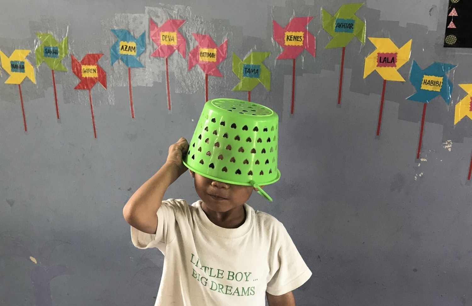 A young child at a daycare wearing a bright green colander as a hat and a t-shirt reading Little Boy Big Dreams, in front of a wall of colourful named paper pinwheels
